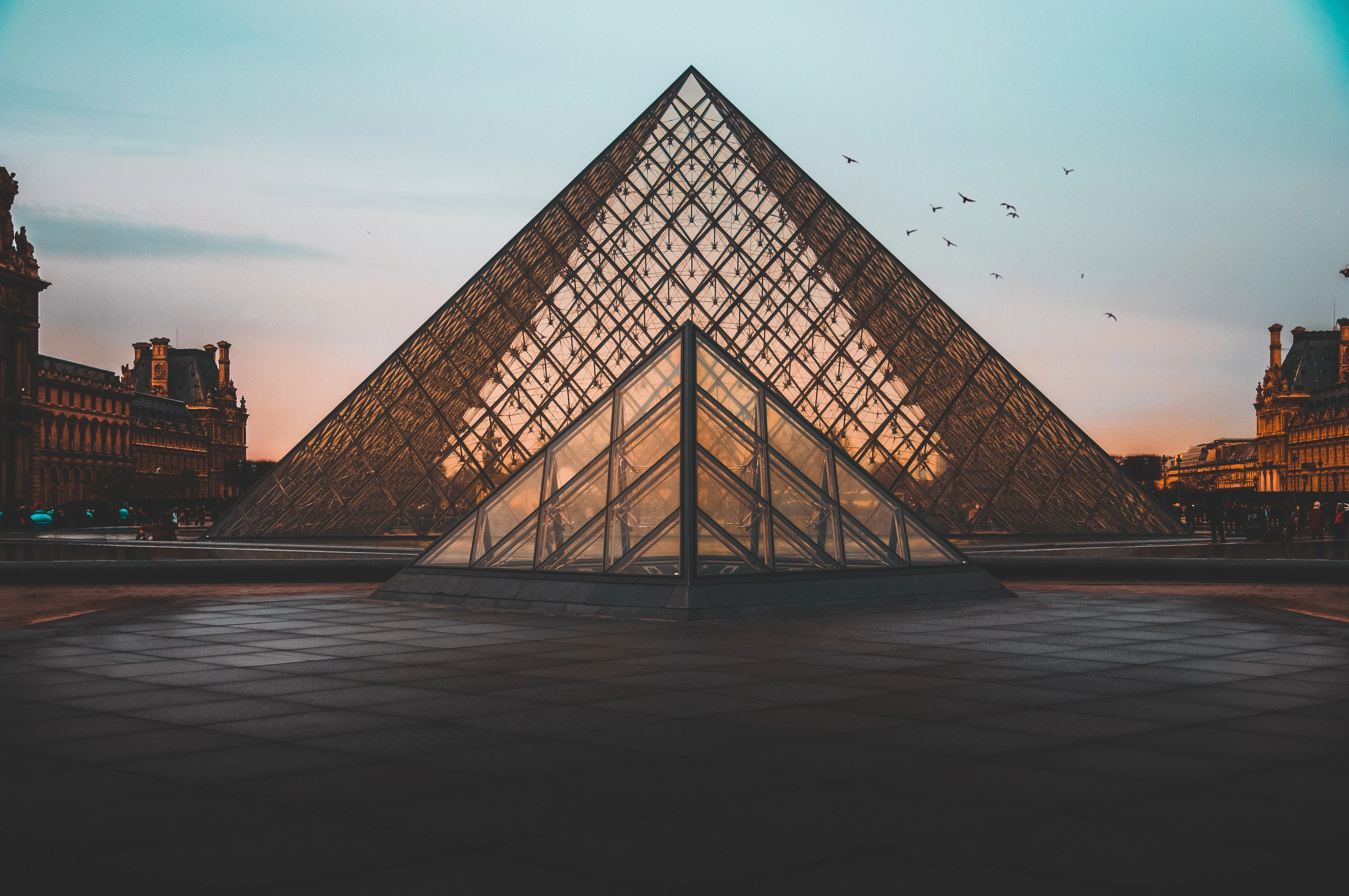 The Louvre Museum silhouetted against a vibrant sunset sky, with warm hues reflecting off its iconic glass pyramid.